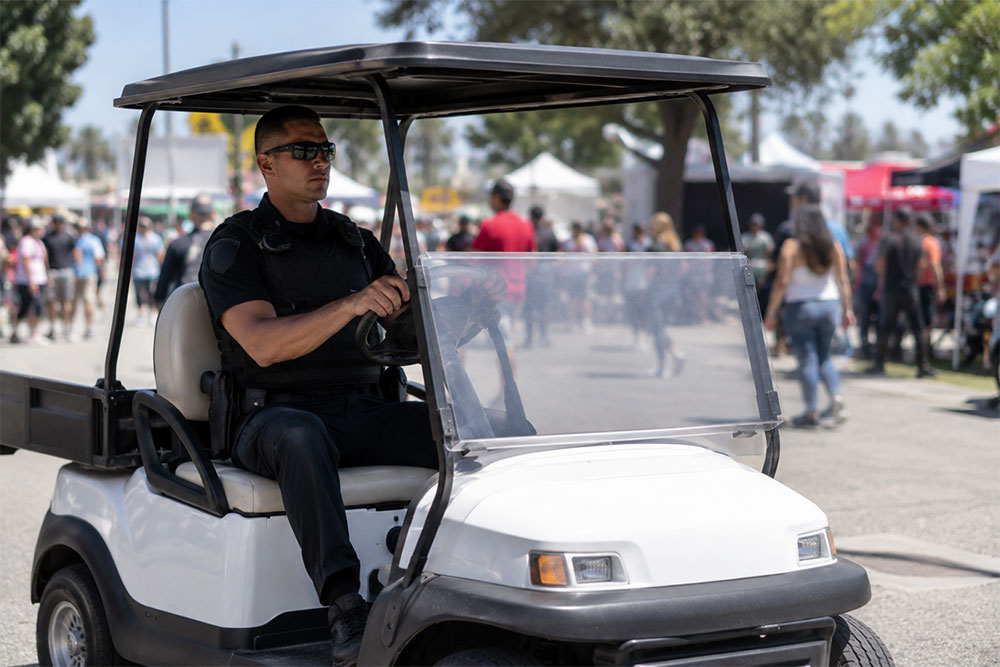 Uniformed security officer driving a golf car through a large outdoor event venue.